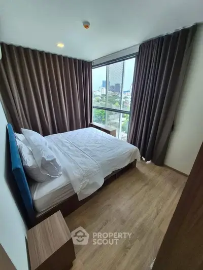 Modern bedroom with large window offering city view, featuring elegant curtains and wooden flooring.