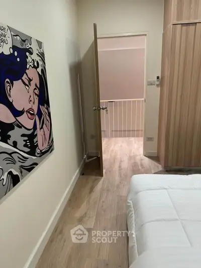 Modern bedroom with stylish wall art and wooden flooring, featuring a cozy bed and open door to hallway.