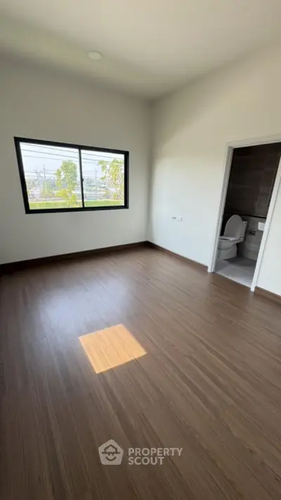 Spacious empty room with wooden flooring and large window, adjacent to a modern bathroom.