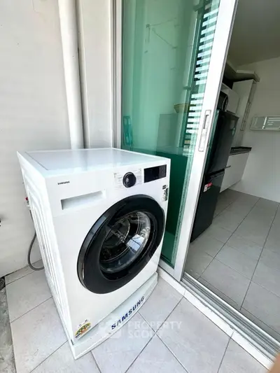Modern apartment with Samsung washing machine on tiled balcony, sliding glass doors to kitchen.