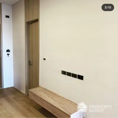 Modern minimalist entryway with sleek wooden bench and matching door, complemented by elegant light switches on a clean white wall.