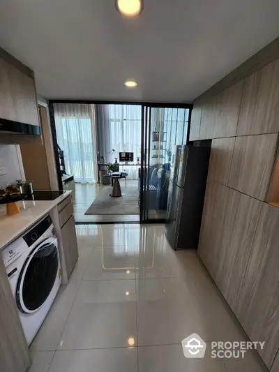 Modern kitchen with sleek cabinetry and washing machine, leading to a stylish living room with floor-to-ceiling windows.