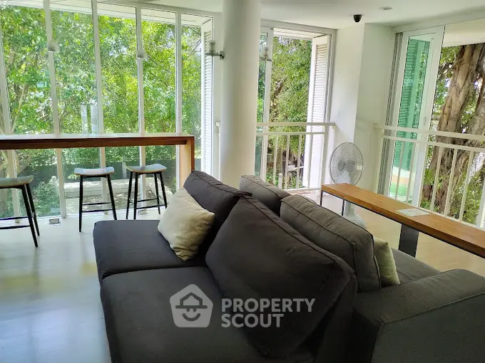 Spacious living room with large windows and lush green view, featuring modern furniture and natural light.