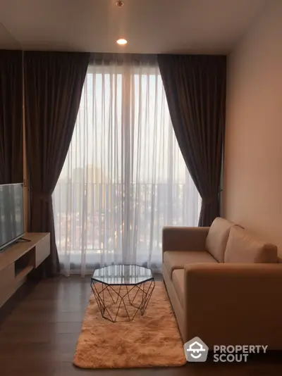 Cozy living room with plush sofa and modern glass coffee table, bathed in natural light from floor-to-ceiling sheer curtains, offering a serene cityscape view.