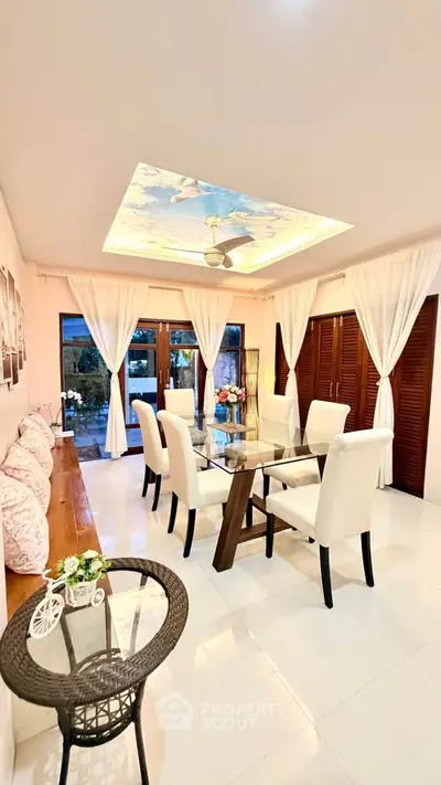 Elegant dining room with glass table, white chairs, and ceiling fan, featuring large windows and stylish decor.