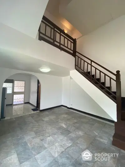 Spacious living area with elegant marble flooring and a grand wooden staircase leading to an upper level, featuring ample natural light and a unique arched doorway.