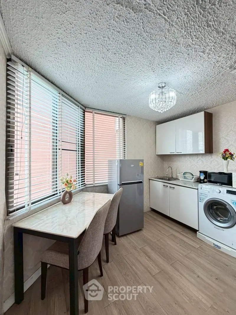 Modern kitchen with dining area, featuring washing machine and fridge, bright natural light from large windows.
