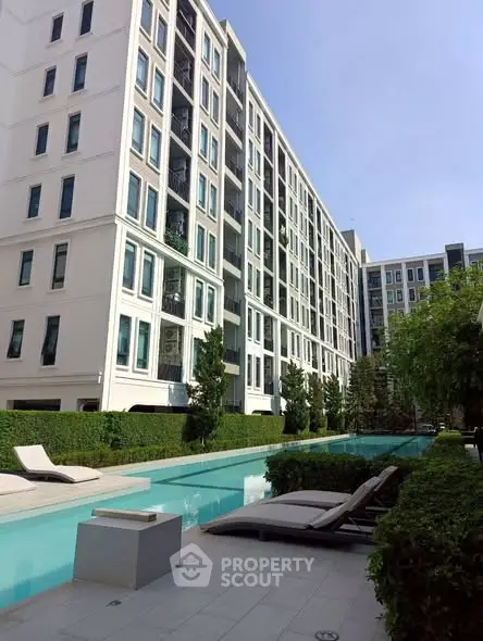 Modern apartment building with luxurious pool and sun loungers in lush garden setting.
