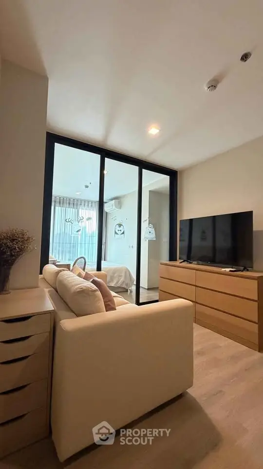 Modern living room with sleek furniture and large TV, leading to a cozy bedroom with glass partition.