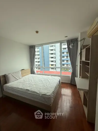 Spacious bedroom with large window offering city view, featuring modern furniture and wooden flooring.