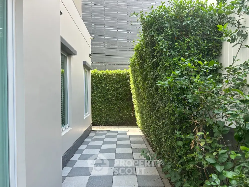 Charming private patio with checkered flooring and lush green hedges, offering a serene outdoor retreat adjacent to a modern home.