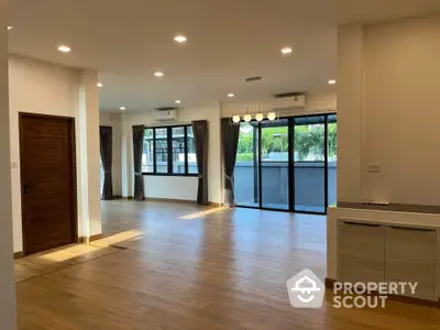 Spacious living room with hardwood floors, ample natural light, and modern lighting fixtures, leading to a cozy balcony with a serene view.