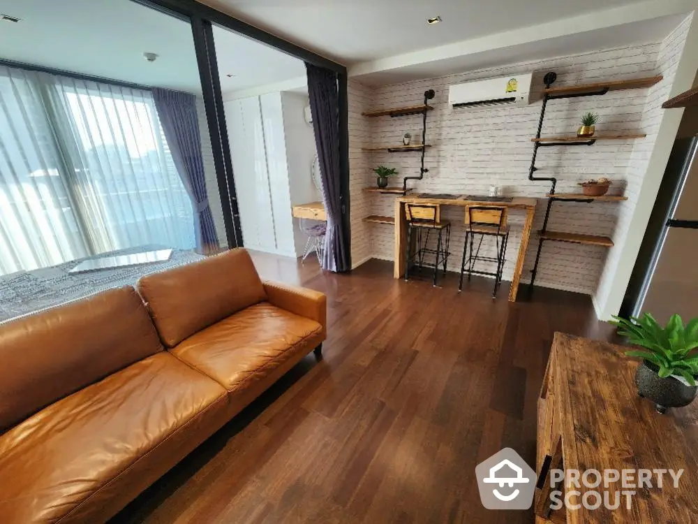 Spacious living room with elegant hardwood flooring, a plush leather sofa, and chic brick accent wall, leading to a sunlit balcony.