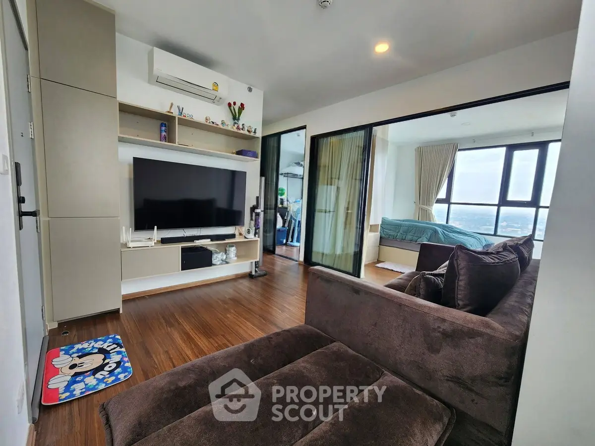 Modern living room with cozy brown sofa and large TV, open to a bedroom with city view.