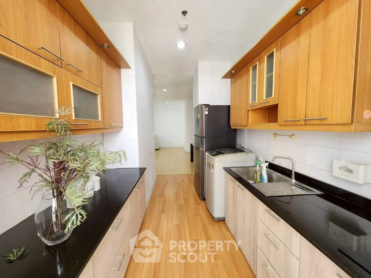 Modern kitchen with sleek black countertops and wooden cabinets, featuring a washing machine and fridge.