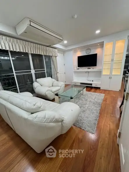 Spacious living room with elegant white furniture and wooden flooring, featuring a large TV and glass coffee table.