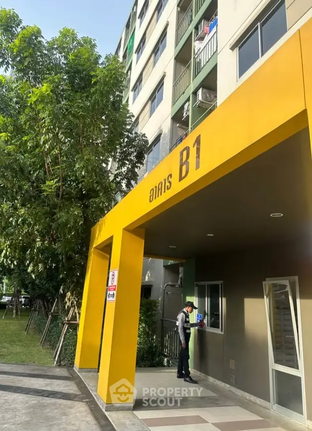 Modern apartment building entrance with vibrant yellow facade and lush greenery.