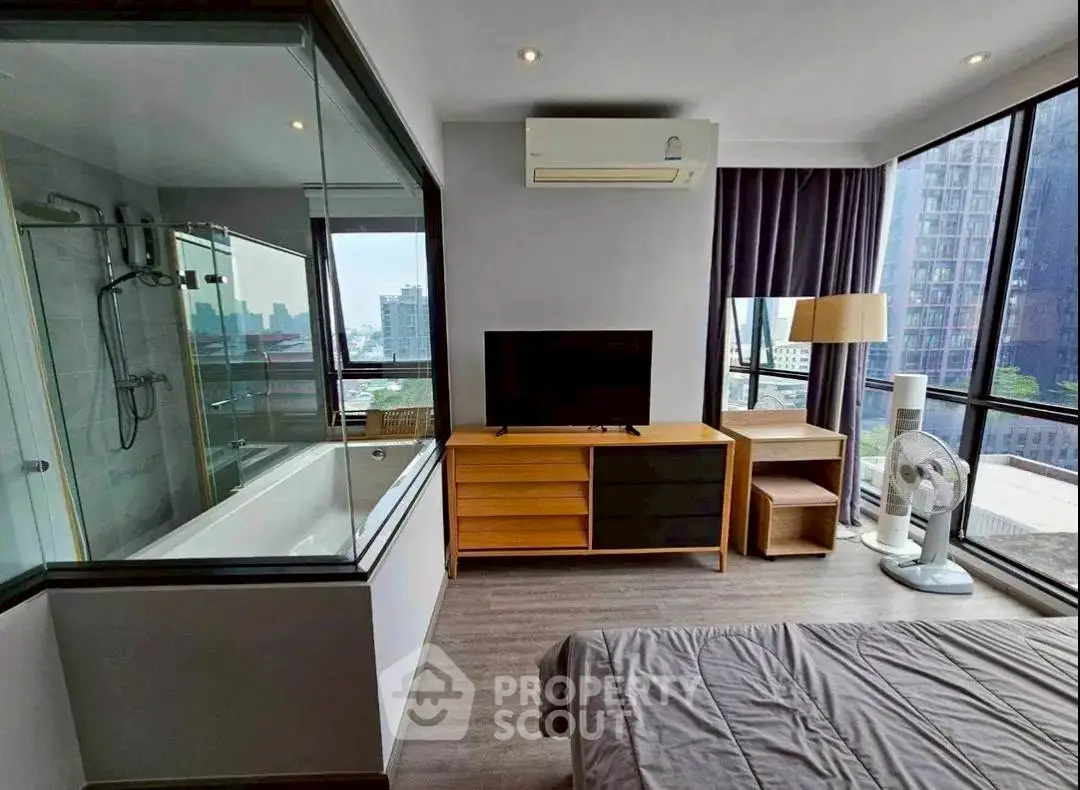 Modern bedroom with city view, featuring glass-enclosed bathtub and sleek furnishings.
