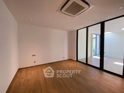 Spacious empty room with wooden flooring and large glass doors leading to a sunlit area.