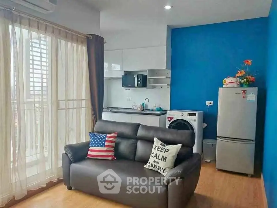 Modern living room with cozy sofa, vibrant blue accent wall, and compact kitchen area featuring appliances.