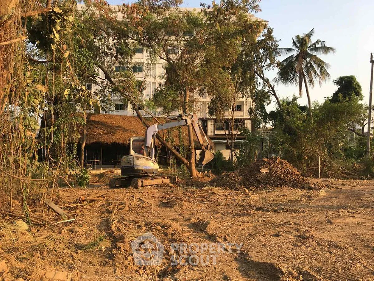 Construction site with excavator and building in background, surrounded by trees and dirt.