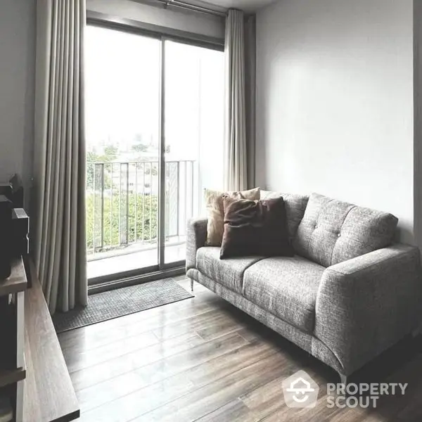 Modern minimalist living room with a comfortable grey sofa, sleek hardwood floors, and access to a cozy balcony with a refreshing view.