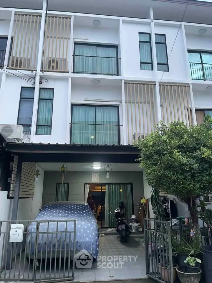Modern three-story townhouse with sleek facade and gated entrance, featuring a small patio and lush greenery.