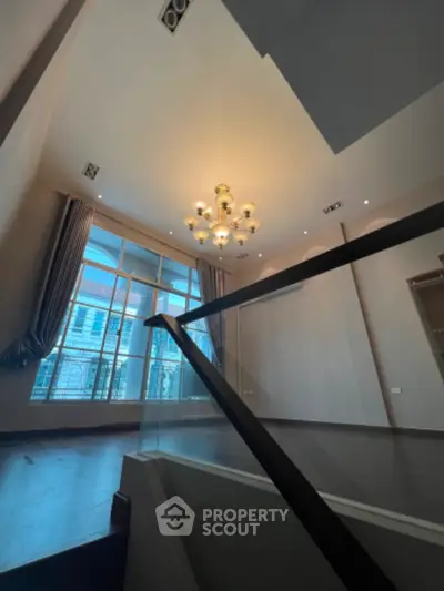 Spacious living room with high ceiling, modern chandelier, large windows allowing ample natural light, and elegant staircase leading to the upper level.