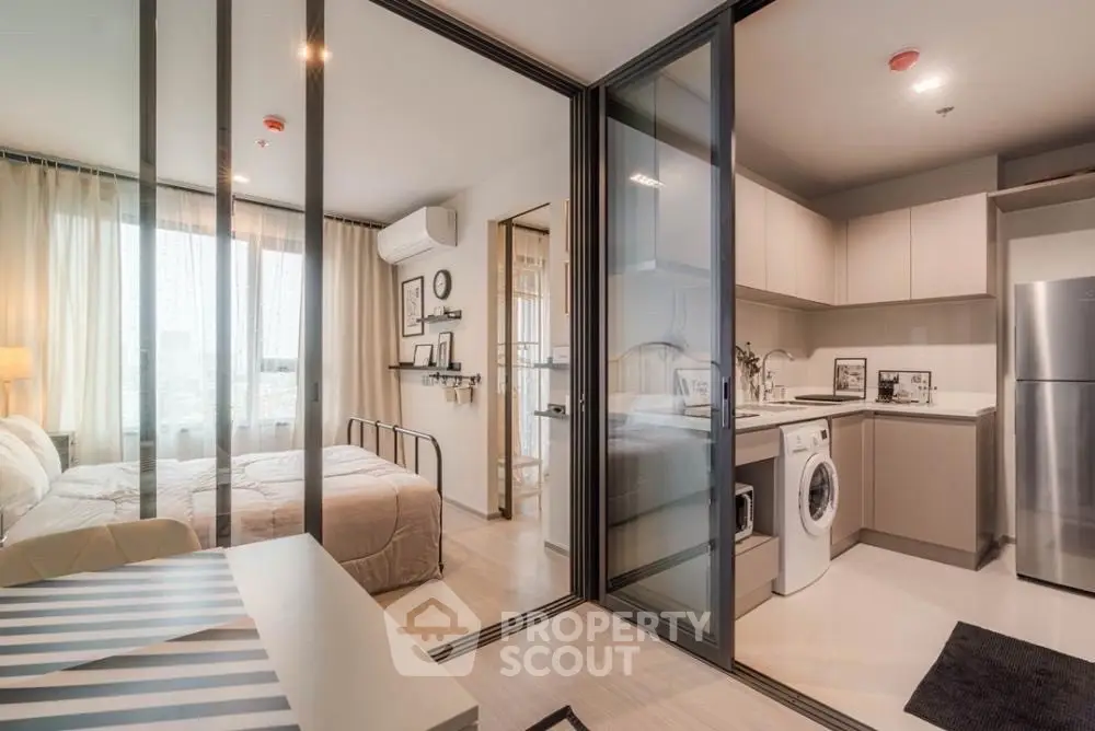 Modern studio apartment with glass partitions, featuring a cozy bedroom and sleek kitchen with washing machine.