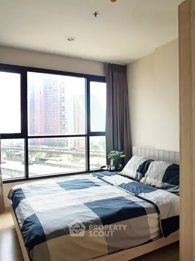 Modern bedroom with large window offering city view, featuring stylish bedding and natural light.