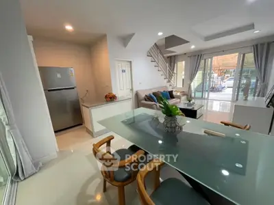 Modern open-plan living room with glass dining table and cozy seating area, featuring a sleek staircase and large windows.