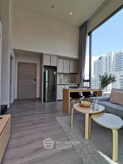 Modern living room with open kitchen and city view, featuring sleek furniture and large windows.