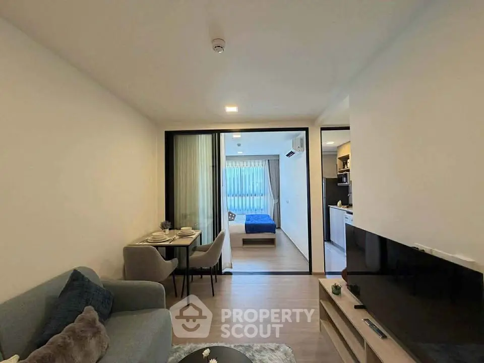 Modern open-plan living room with dining area and view into bedroom, featuring sleek furniture and natural light.
