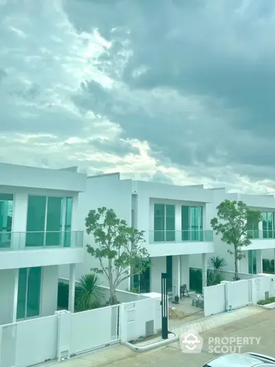 Modern white townhouse complex with large windows and lush greenery under a cloudy sky.