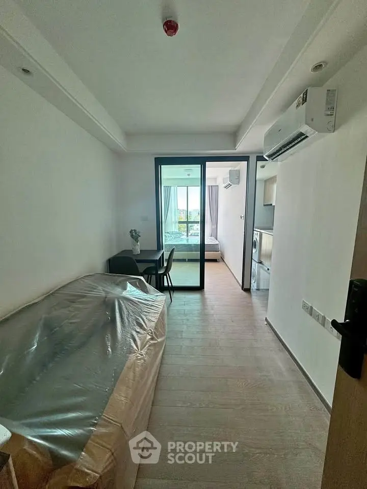Modern apartment interior with open layout, featuring a compact living area and kitchen, leading to a bright bedroom.