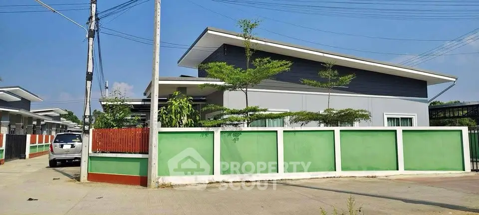 Modern single-story house with unique slanted roof and green fence in sunny neighborhood.