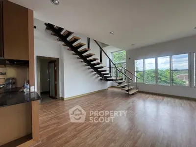 Spacious modern living room with wooden flooring and stylish staircase, featuring large windows with lush green views.
