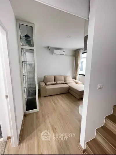 Modern living room with beige sectional sofa and wooden flooring, featuring a sleek glass cabinet.