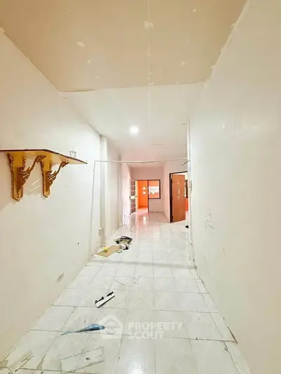 Spacious hallway with tiled flooring and wall-mounted shelf, leading to a bright room.