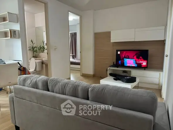 Modern living room with gray sofa and wall-mounted TV, open view to bedroom and bathroom.