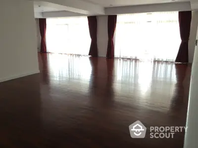 Spacious empty living room with polished wooden floors and large windows with curtains.