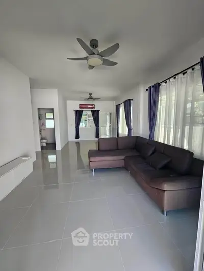 Spacious living room with modern sofa and ceiling fan, large windows with curtains, and sleek tiled flooring.