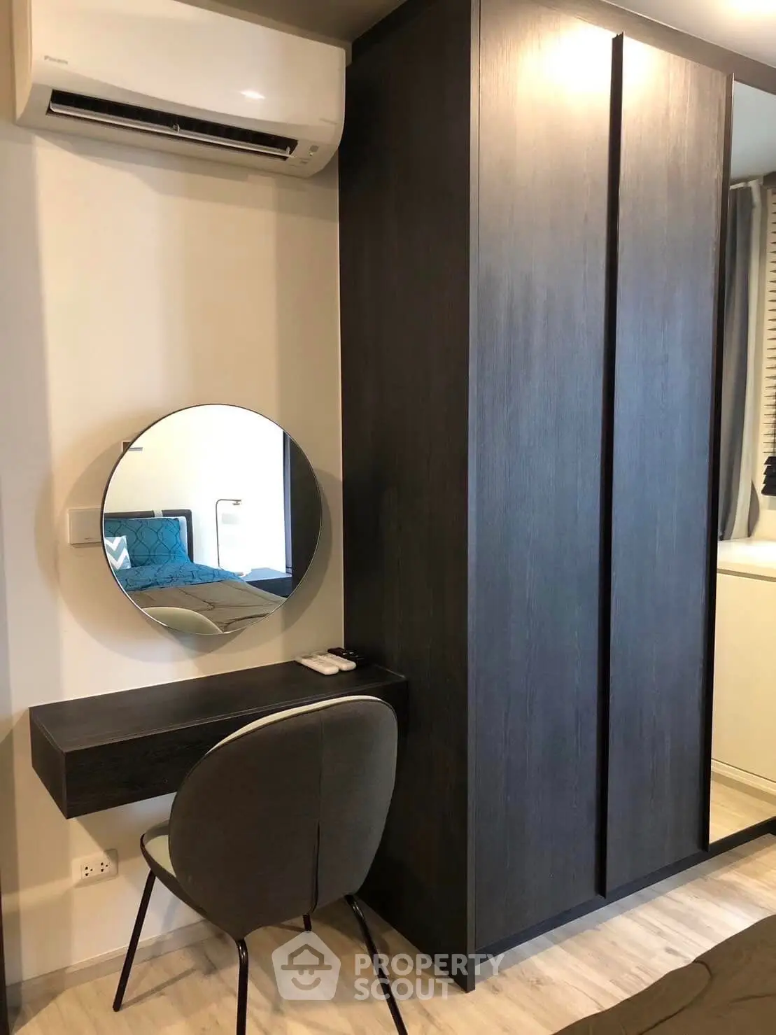 Modern minimalist bedroom with sleek dark wood wardrobe, stylish round mirror, and cozy study nook, complemented by a comfortable chair and efficient air conditioning.