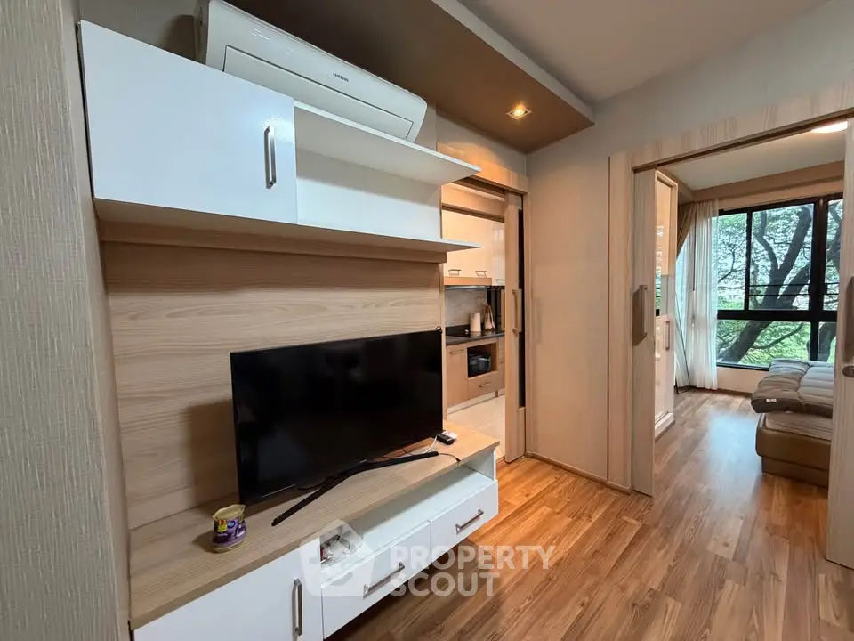 Modern living room with wooden flooring and stylish TV unit, leading to a cozy bedroom with large windows.