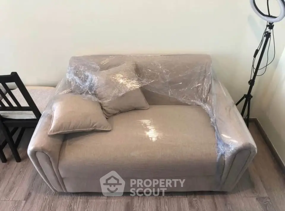 Cozy beige sofa wrapped in plastic with pillows, next to a black chair and ring light on wooden floor.