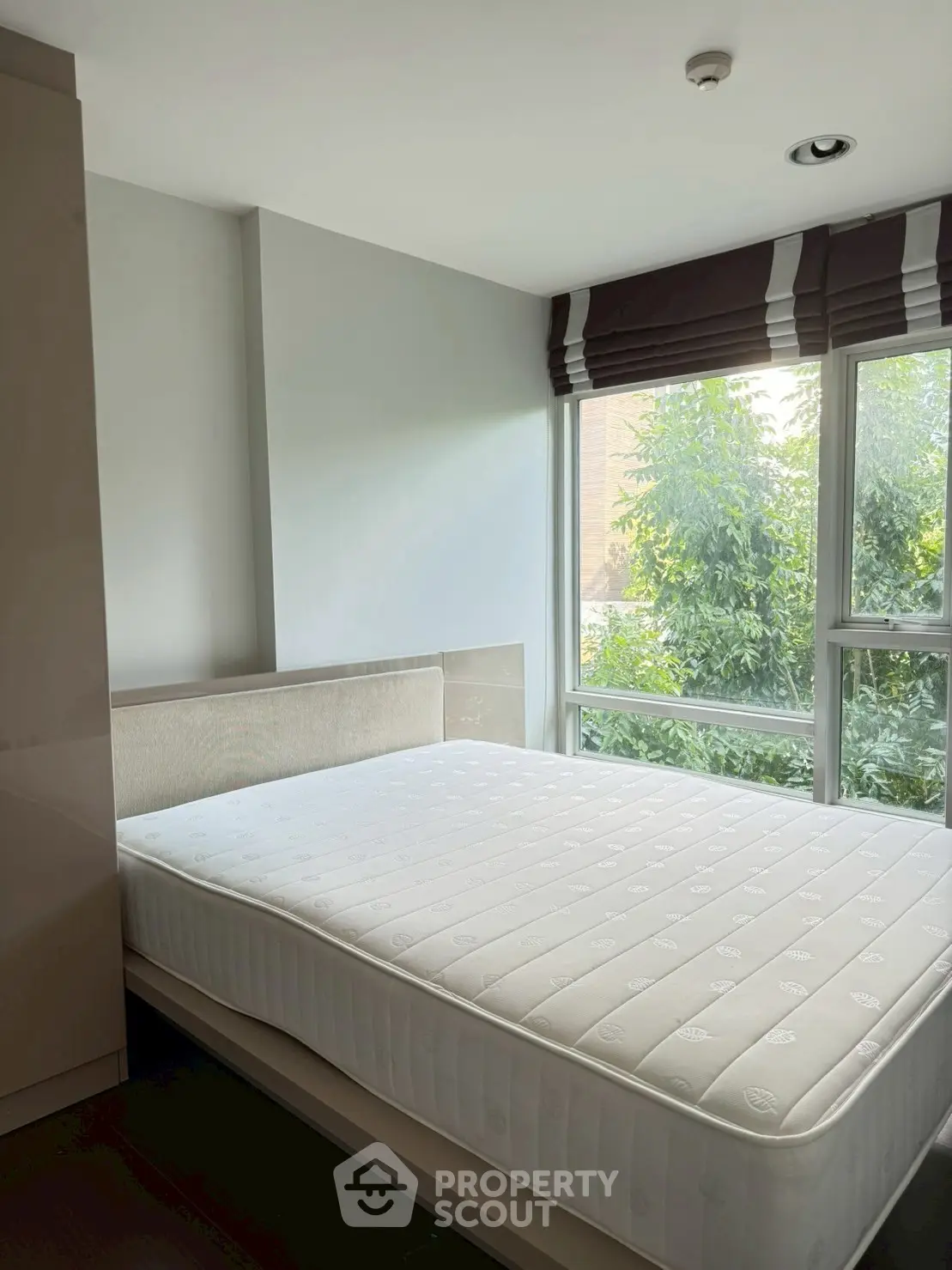 Bright bedroom with large window and comfortable mattress, offering a serene view of lush greenery.