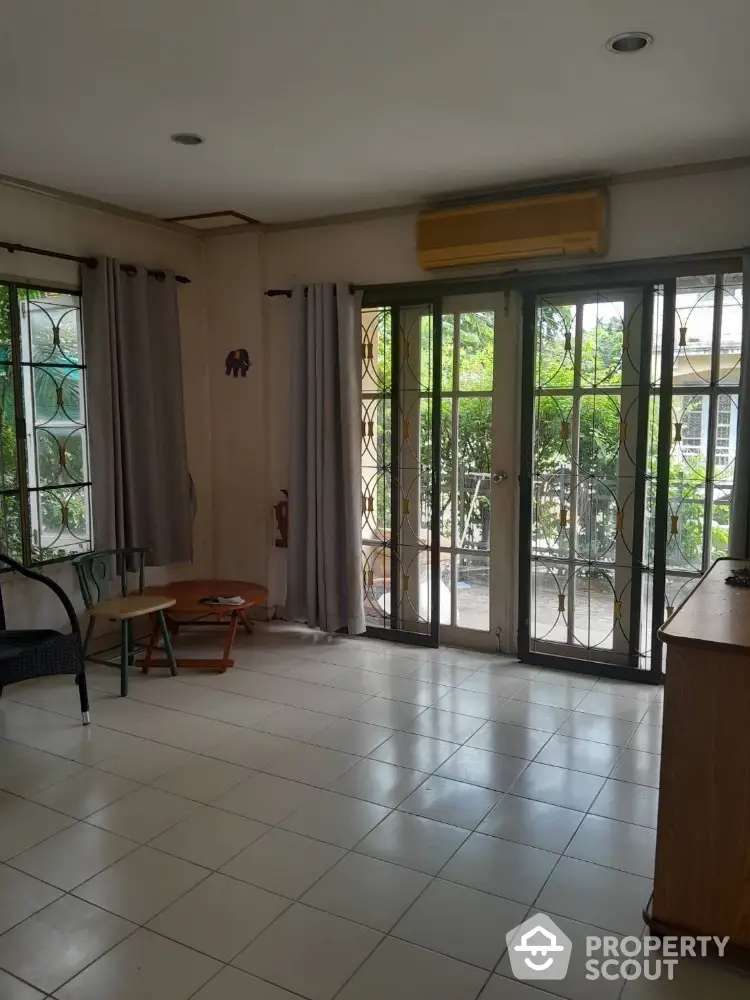 Spacious living room with abundant natural light, featuring large windows, tiled floors, and a serene garden view, perfect for comfortable living and entertaining guests.