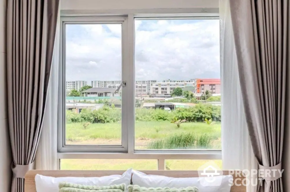 Bright bedroom with large window offering scenic view of greenery and modern buildings.