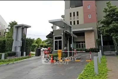 Modern gated entrance of residential building with security booth and lush greenery.
