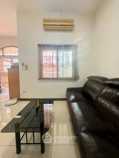 Modern living room with leather sofa and glass coffee table, featuring air conditioning and natural light.